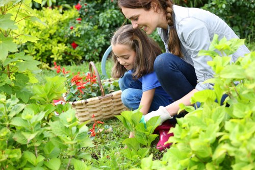 In a lush outdoor garden scene, a woman and a young girl are engaged in gardening activities amidst vibrant green foliage, flowering plants, and a well-maintained lawn. The woman, dressed casually with her hair in a braid, is smiling as she supervises the girl, who is focused and wearing a blue shirt. They are surrounded by various garden plants, including flowering bushes with red blossoms, and are working near a patch of grass and soil. The garden features neatly trimmed hedges, decorative flower beds, and a small pathway with paving stones. Natural sunlight illuminates the scene, casting gentle shadows and highlighting the healthy, dense greenery typical of a landscaped outdoor space in Shoreditch. This image exemplifies outdoor gardening and landscape maintenance efforts, supported by professional services from Landscapers Shoreditch, aligning with the themes of lawn care, planting, and garden upkeep that enhance residential outdoor areas through expert landscaping.