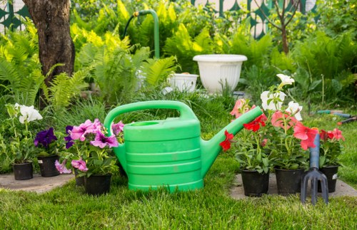 The image depicts a garden scene with a neatly maintained grassy lawn in the foreground, bordered by various potted flowering plants in shades of purple, white, and pink. A central green plastic water can is prominently placed among the plants, and nearby is a small gardening fork with a red handle. In the background, lush green foliage and fern-like plants are visible, along with a few larger shrubs and a tree trunk, creating a natural, vibrant outdoor environment typical of a private garden in Shoreditch. The scene suggests a well-tended outdoor space that could benefit from ongoing gardening and landscaping services by companies such as landscapersshoreditch.co.uk, integrating functional gardening tools and colorful plant displays. The lighting appears natural, indicating a mild weather day, enhancing the lush and healthy appearance of the garden plants and grass.