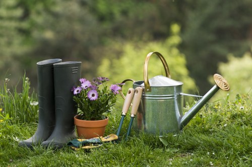 A garden scene showing a patch of well-maintained lawn with lush, green grass in the foreground, bordered by garden tools including a pair of gardening shears, a small trowel, and a rake. Nearby, there is a terracotta pot filled with purple and white flowering plants, adding colour to the scene. To the right, a silver metal watering can with a wooden handle is positioned on the grass. Behind these items, a pair of black rubber gardening boots stands upright, alongside a wooden garden fence and blurred trees in the background, indicating an outdoor space typical of a Shoreditch garden. The environment appears to be a well-kept backyard or front garden area suitable for outdoor gardening projects. The weather looks clear, suggesting a bright, sunny day, ideal for planting or maintaining flower beds. This scene illustrates typical gardening activity, supporting professional gardening services focused on planting, lawn care, and outdoor maintenance around Shoreditch or similar urban areas.