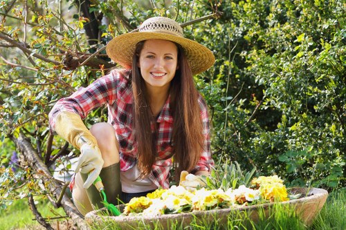 A young woman with long brown hair, wearing a straw hat, red and white plaid shirt, and gardening gloves, is crouching in a lush garden. She is smiling and tending to a flower bed filled with blooming yellow and white flowers, which is bordered by a wooden edging. Behind her, there are dense green hedges, small trees, and various plants, indicating a well-maintained landscaped yard. The grass is vibrant and neatly trimmed, and the overall environment looks bright with natural sunlight filtering through the foliage. The scene exemplifies typical outdoor gardening activities in a residential garden, aligning with professional gardening and landscaping services offered by Landscapers Shoreditch near the London area, possibly within the E15 postcode district.