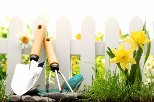 The image depicts a small garden scene in a residential outdoor space, with a white picket fence in the background that suggests a suburban or urban environment near Shoreditch. In the foreground, there is a patch of well-maintained, lush green grass with dense, healthy blades. To the right, a cluster of bright yellow daffodils with green leaves adds vibrant colour and natural contrast to the scene. Slightly to the left, a few white daisies with yellow centres are visible, providing variety in plant types and natural textures. Resting on the soil and grass near the fence are gardening tools including a small trowel with a wooden handle, a hand rake, and a pair of garden gloves, indicating recent or ongoing outdoor work. In the background, the garden appears to be part of a neat and tidy outdoor space possibly used for leisure or small-scale planting. The overall environment is well-lit with natural daylight, suggesting a clear day, and the scene aligns with gardening and landscaping services such as lawn care, planting, and garden maintenance offered by companies like Landscapers Shoreditch in the local area.