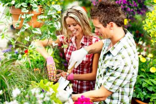 The image depicts a young man and woman working together in a lush, well-maintained garden with a variety of plants, shrubs, and flowering borders. The garden features a central lawn area with dense, green grass that appears carefully mowed, bordered by vibrant flower beds filled with colorful blooms and leafy foliage. The background includes a wooden lattice structure supporting climbing plants, along with a natural mix of flowering bushes and small trees providing depth to the outdoor space. The individuals are wearing gardening gloves and casual clothing suitable for outdoor work, actively engaging with garden tools to tend to the plants. Soft natural light highlights the vivid green hues and vibrant flower colours, suggesting a bright, sunny day. This scene reflects typical elements of professional gardening services, combining lawn maintenance, planting, and outdoor beautification in a suburban residential setting, possibly in Shoreditch or nearby areas, supporting local SEO relevance for gardening and landscaping services in the region.