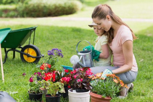 In a well-maintained garden with a lush, green lawn typical of the Shoreditch area, a woman and a young girl are engaged in gardening activities. They are tending to a variety of potted plants, including vibrant flowering species with red, pink, purple, and orange blooms, placed on the grass. The garden features a border of hedges in the background, with a paved patio area partially visible behind them. Nearby, a wheelbarrow with a yellow wheel is positioned on the grass, indicating ongoing outdoor work. The scene is set during a bright, clear day with natural sunlight illuminating the garden space, emphasizing the natural tones of the plants and lawn. The overall environment suggests a landscaped backyard or front garden designed for leisure and gardening, supported by professional outdoor maintenance, such as by Landscapers Shoreditch, to enhance the outdoor comfort and visual appeal of properties in the area.