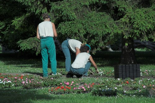 The image depicts three individuals working in a lush, well-maintained garden with dense green foliage and mature trees creating a shaded backdrop. Two of them are bent over, tending to a flower bed bordered with a variety of small, colorful flowering plants, while the third person stands nearby, observing or waiting to assist. The flower bed features a mixture of pink, red, and white blossoms, contrasting with the dark, rich soil and bright green leaves. The surrounding lawn is neatly trimmed, with vibrant, healthy grass covering the foreground, and a section of black garden edging or screening can be seen on the right side of the image. The scene appears to be a peaceful outdoor space on a clear day with natural light filtering through the canopy, exemplifying typical garden maintenance work that could be performed by professional landscapers or gardening services in Shoreditch or the surrounding areas, focusing on flower bed planting and lawn care.