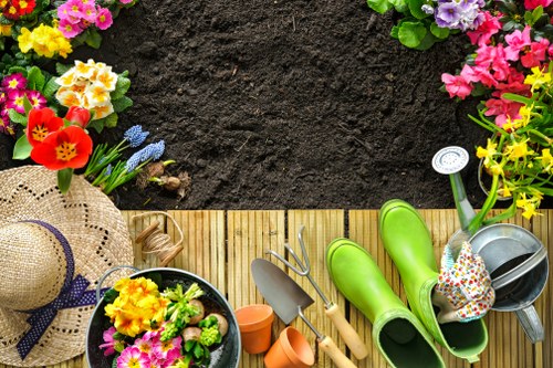 The image shows a neatly arranged back garden space with a wooden decking area bordered by vibrant flower beds containing colorful blooms such as pink, yellow, and purple flowers. In the foreground, gardening tools including a trowel and watering can are placed on the decking alongside a pair of bright green waterproof garden boots, indicating recent or ongoing planting work. A straw sun hat with a black ribbon is positioned on the left side, resting near a black plastic pot filled with freshly planted flowers. Behind the flower beds, there is a patch of dark, well-tilled soil, ready for planting or recent planting activity. The outdoor environment appears well-maintained and inviting, suitable for gardening and landscaping services offered by Landscapers Shoreditch in the area of Shoreditch, London, with natural lighting suggesting daytime conditions.