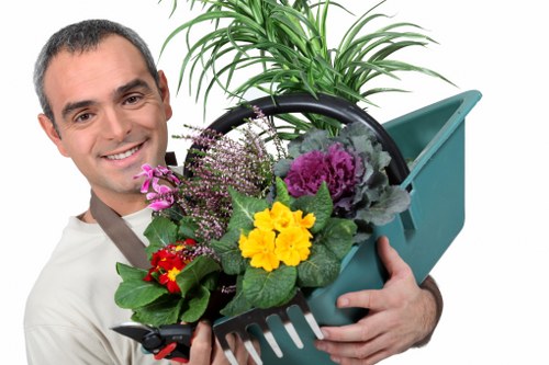 The image depicts a man with short dark hair and a friendly expression, holding a small plastic garden trowel and a black container filled with a variety of plants and flowers, including purple, yellow, and red blooms, set against a plain white background. The plants appear healthy with vibrant green leaves and colorful flowers, suggesting they are recently purchased or ready for planting. The tools and plants imply a focus on gardening activities such as planting or garden maintenance. Although not part of the outdoor environment, the image conveys a gardening theme that aligns with professional landscaping and garden care services offered by companies like Landscapers Shoreditch in the London area. The overall scene emphasizes garden enhancement, planting, and outdoor maintenance work, subtly supporting local SEO for gardening services in Shoreditch or nearby postcode areas, with a friendly, professional tone suitable for a gardening service website.