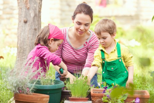 A young woman with dark hair pulled back, wearing a pink and white striped long-sleeve top, is gardening outdoors in a landscaped backyard. She is assisting a little girl in a pink headband and a purple jacket, as they tend to potted plants on a garden table, with the girl using a small gardening tool. Beside them, a young boy with blonde hair, dressed in a yellow top and green overalls, observes and participates in the activity. The garden setting features a grassy lawn with lush, healthy plants and a background of tall trees and a wooden fence, creating a natural and inviting outdoor environment. Potted herbs and flowering plants are arranged on the table, with some in round ceramic containers and others in rectangular flowerbeds. The scene is illuminated by soft natural daylight, suggesting a mild, pleasant weather suitable for outdoor gardening activities, supporting gardening and landscaping services in Shoreditch and nearby areas.