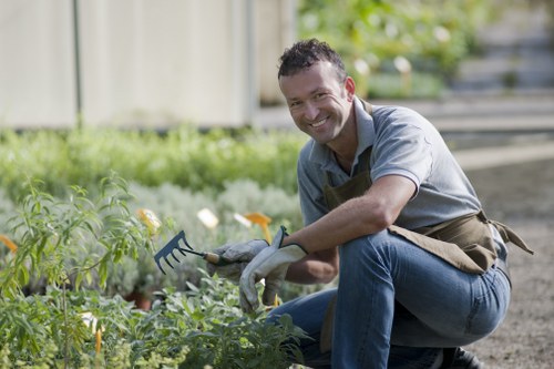 The image shows a man kneeling in a small, well-maintained garden, surrounded by various green plants and flowers with bright yellow blooms. He is holding a small garden rake and wearing gardening gloves, a beige apron, and casual clothing suitable for outdoor work. The garden features a mix of ornamental shrubs and flower beds with dark soil visible between the plants. In the background, a wooden fence and a paved pathway can be seen, indicating a private residential outdoor space. The scene appears to be in bright natural daylight, suggesting a clear weather condition, ideal for gardening activities. This setting exemplifies typical outdoor garden maintenance and landscape care, which a professional gardening service like Landscapers Shoreditch could support, especially in urban areas with small, managed gardens close to Shoreditch.