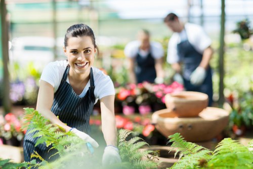 In a well-lit greenhouse setting, a smiling woman wearing a striped gardening apron and gloves tends to lush green ferns and other potted plants in the foreground. Behind her, two other gardeners, dressed in similar attire, are seen working among vibrant flowering plants arranged in containers on tables, with some in the process of planting or pruning. The greenhouse structure features large glass panels that allow natural daylight to illuminate the space, creating a bright and welcoming environment for outdoor gardening and landscaping activities. The background shows a variety of garden elements, including decorative stone water features, wooden plant stands, and neatly arranged flower beds, highlighting the professional care and landscape design typically provided by companies like Landscapers Shoreditch. The scene emphasizes meticulous plant maintenance, outdoor environmental enhancement, and the natural textures of soil, foliage, and paving, all set within an urban garden scheme in the Shoreditch area, which supports high-quality gardening services.